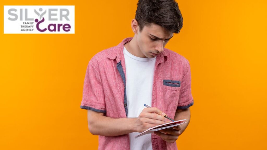 Male teen standing against a yellow background, taking notes in a pad journal, reflecting self-esteem and focus.