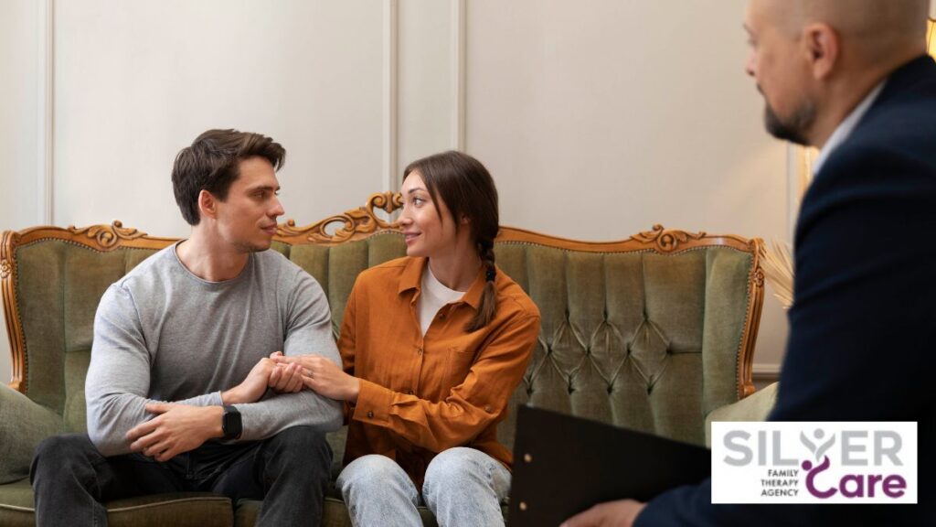 A couple receives therapy while sitting on a coffee-colored sofa, holding each other’s arms and looking at each other.