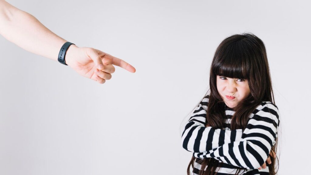 A young child folds her arms and leans against a wall, looking upset while being reprimanded.