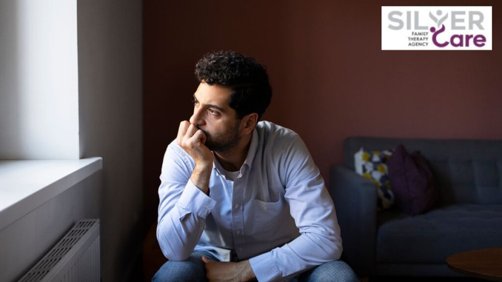 A man in a living room stares out the window, resting his chin on his hands with fingers folded against his lips.