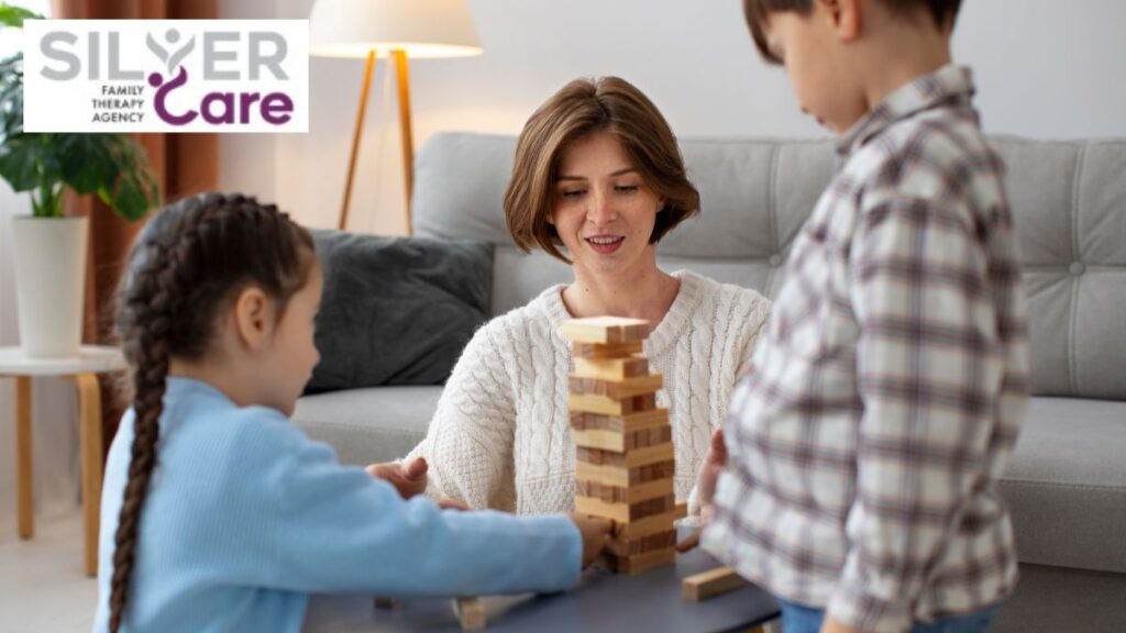 A female therapist guides two children as they stack a tower of blocks on a living room table.